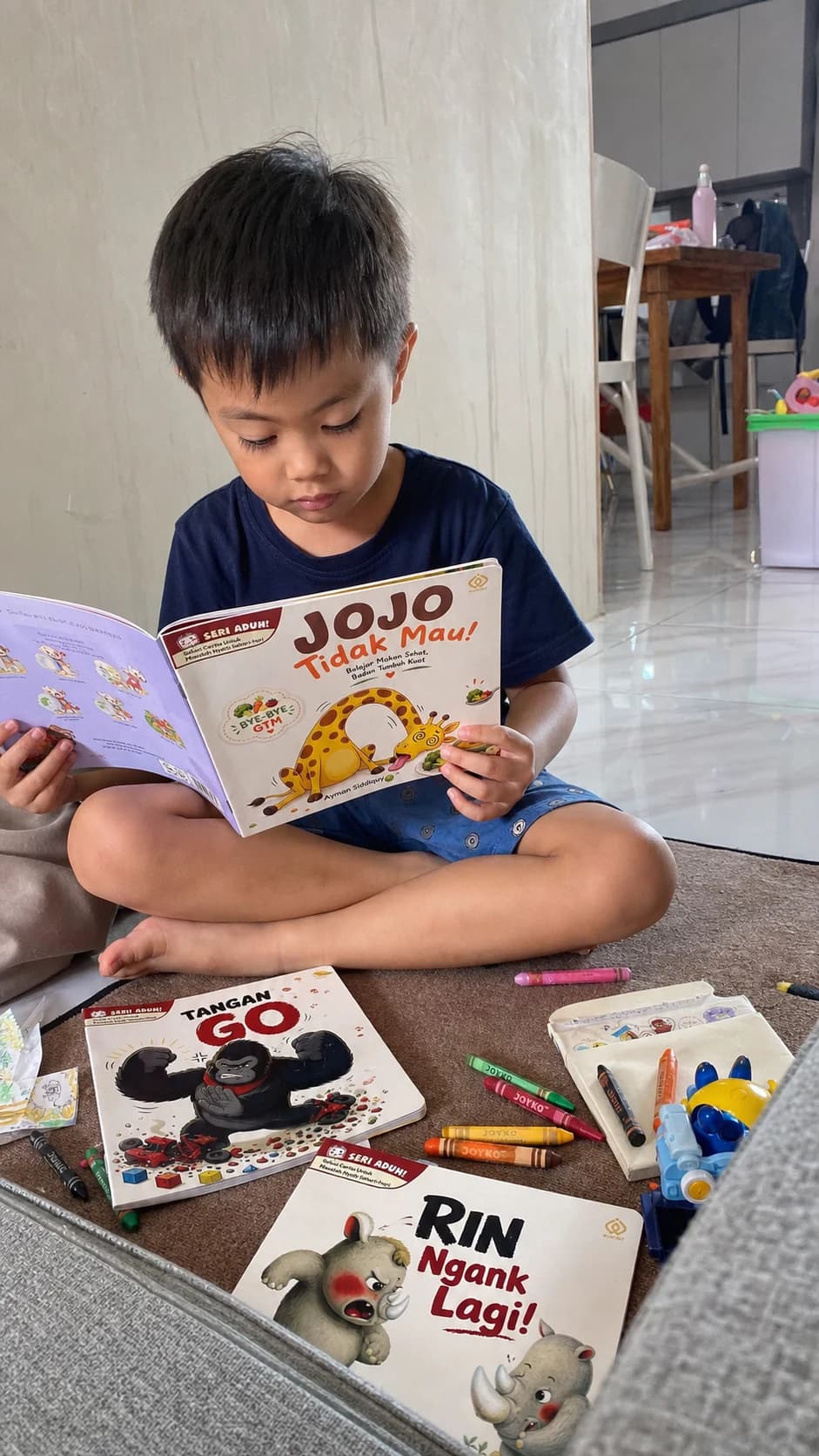 Anak membaca buku Jojo Tidak Mau dengan beberapa buku Seri Aduh di dekatnya